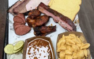 a tray with baked beans, Mac and cheese, brisket, wings, sausage, a rib, two pieces of bread and pickle slices.