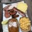 a tray with baked beans, Mac and cheese, brisket, wings, sausage, a rib, two pieces of bread and pickle slices.