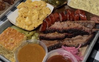 A tray of BBQ food. Brisket, sausage, cornbread, Mac and cheese, collard greens and sauces from Rev BBQ in Rochester, NY.